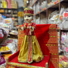 Wooden idol male doll in red-and-gold top and yellow skirt, standing on a red base.