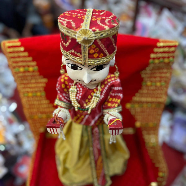 Wooden idol doll with black eyebrows and red crown, red beaded outfit, hands raised.