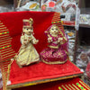 Wooden deity idol figurines pair, seated dolls in gold and magenta dresses on red base.