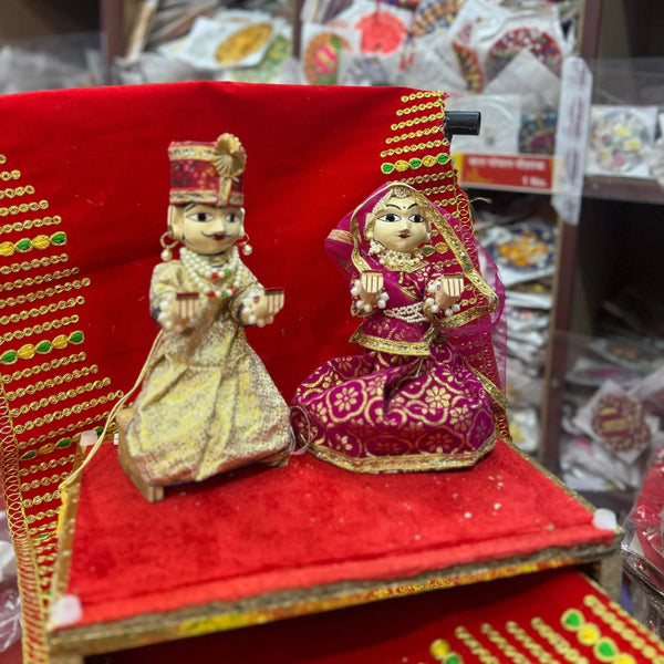 Wooden deity idol figurines pair, seated dolls in gold and magenta dresses on red base.