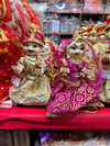 Wooden deity idol figurines, two seated dolls with gold crown and red, magenta patterned dresses.