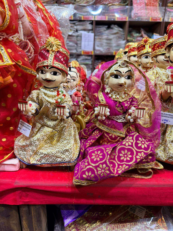 Wooden deity idol figurines, two seated dolls with gold crown and red, magenta patterned dresses.