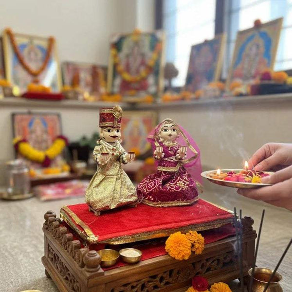Wooden deity idol on carved pedestal, two seated dolls in gold and magenta outfits.